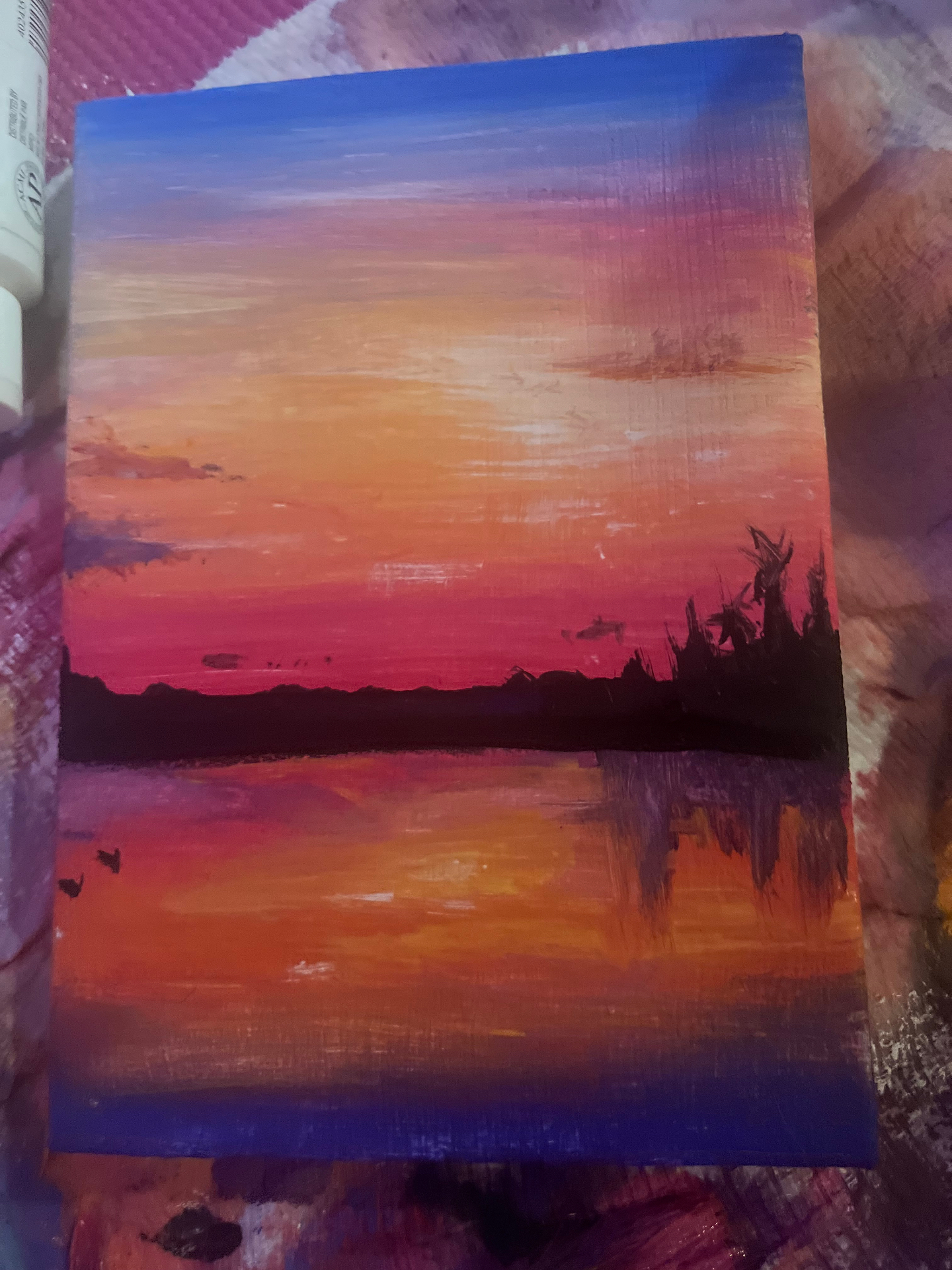 Acrylic painting of an orange and pink sunset reflected over water with a dark treeline